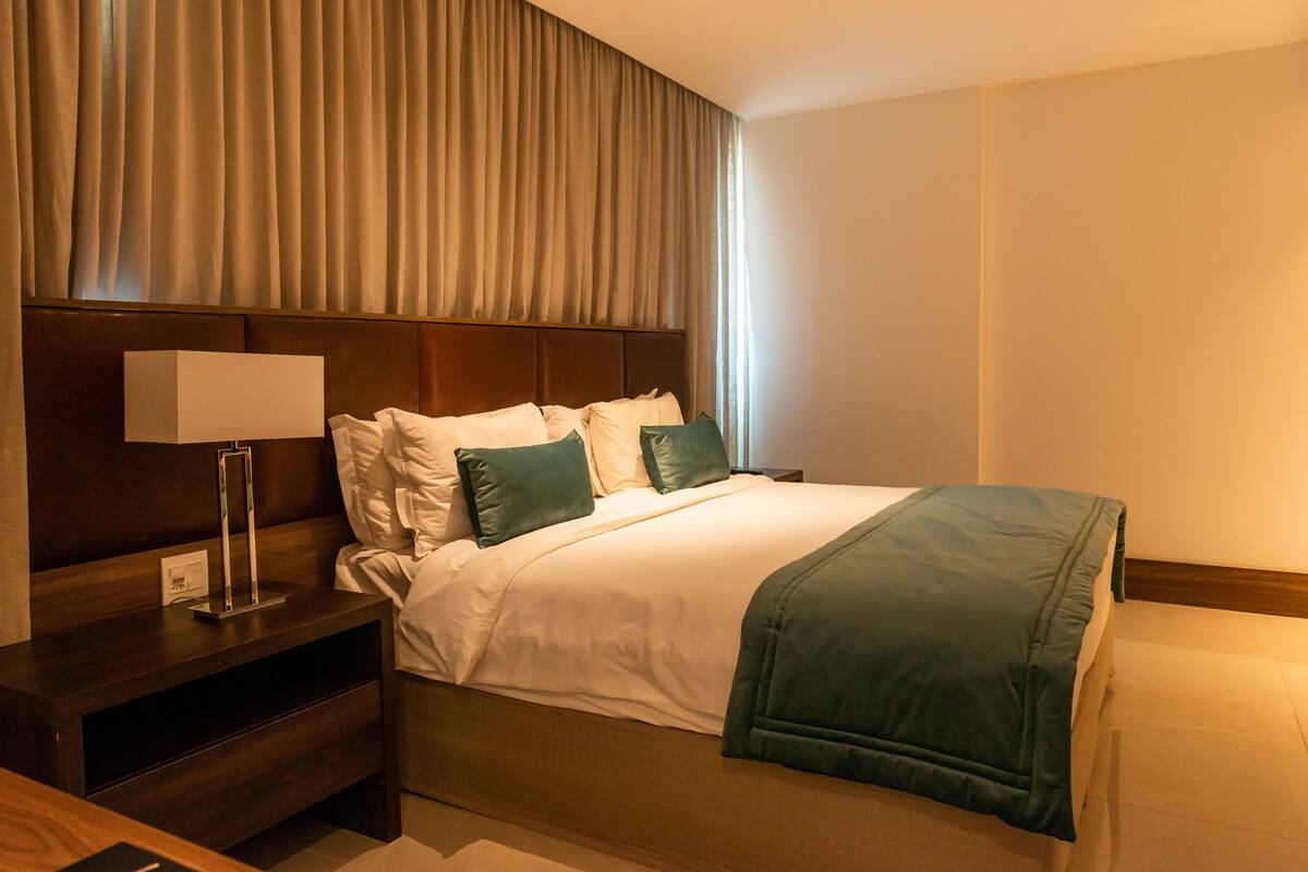 A hotel room with a bed, a nightstand, a lamp, and a window with curtains.