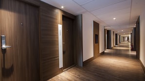 Corredor moderno de hotel com piso e paredes de madeira, com várias portas e um elevador visível.