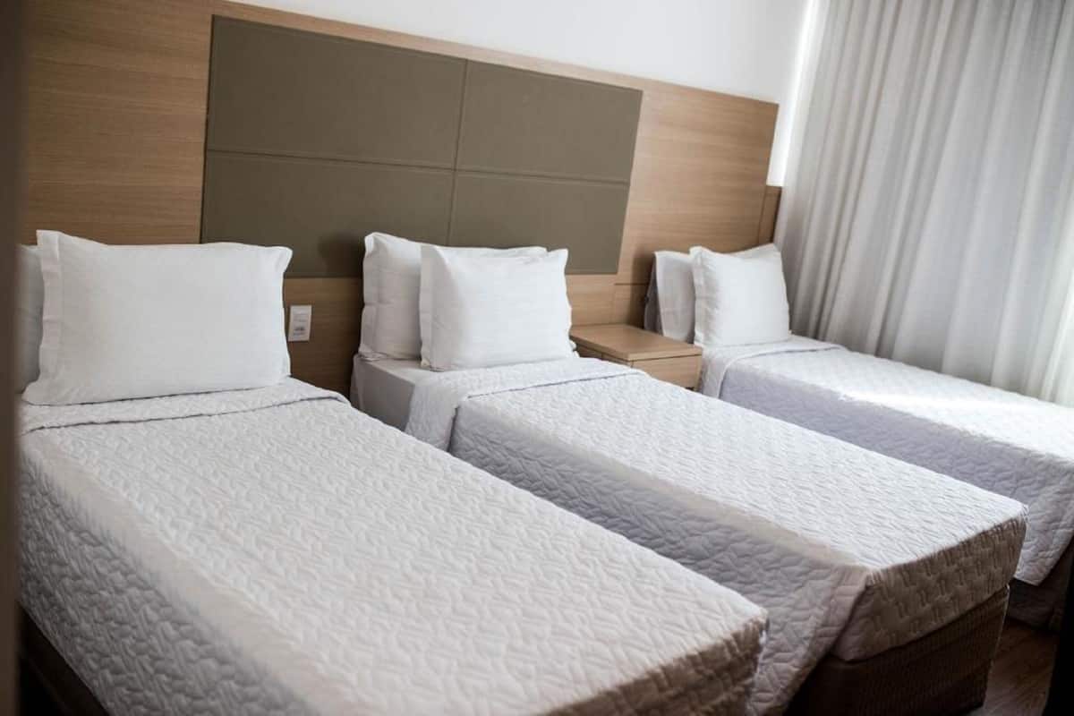 A hotel room with three single beds, each with a white quilted mattress and pillow. The headboard is wooden with a modern design, and there is a window with white curtains.