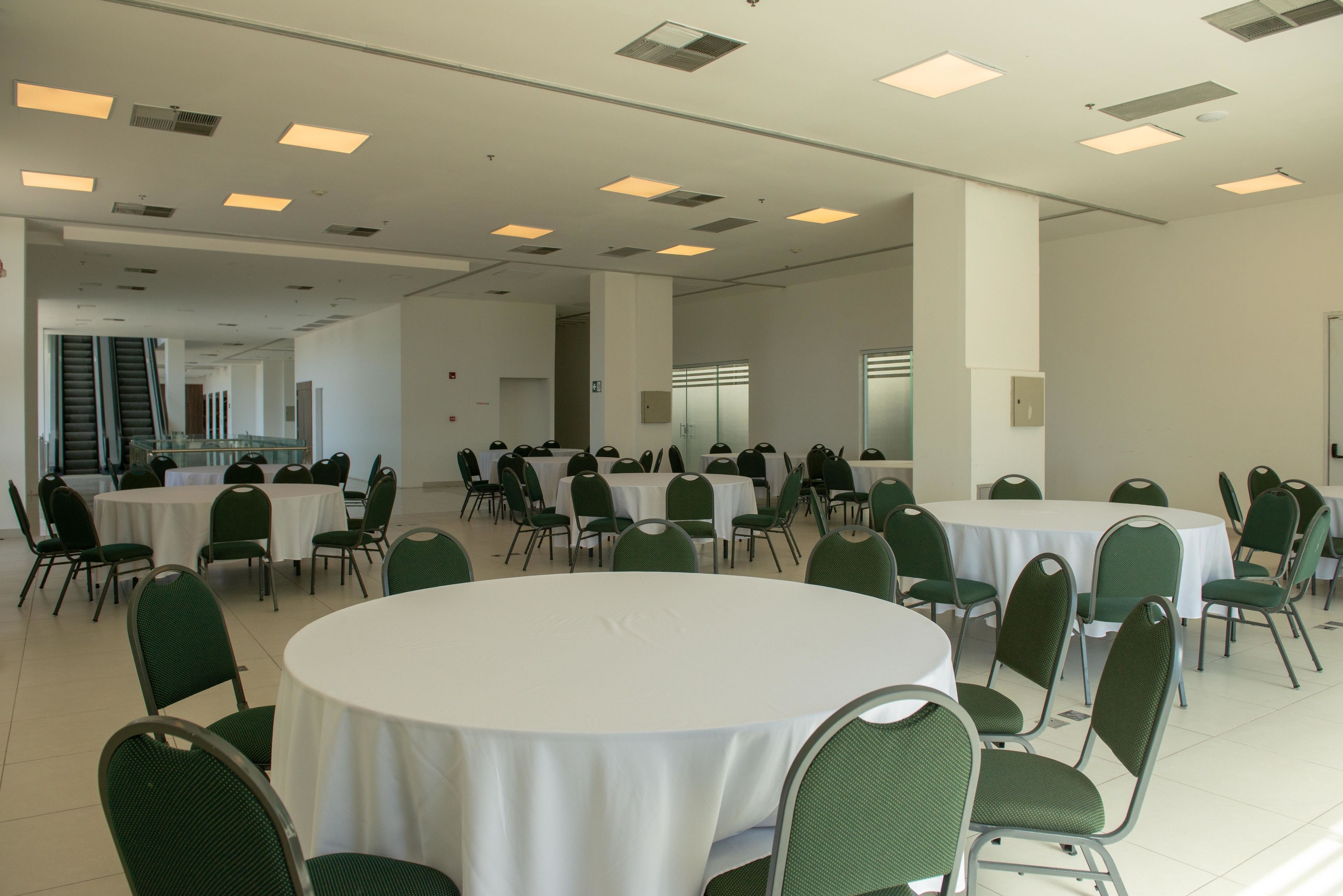 A spacious conference room with multiple round tables and green chairs, equipped with ceiling lights and a tiled floor.