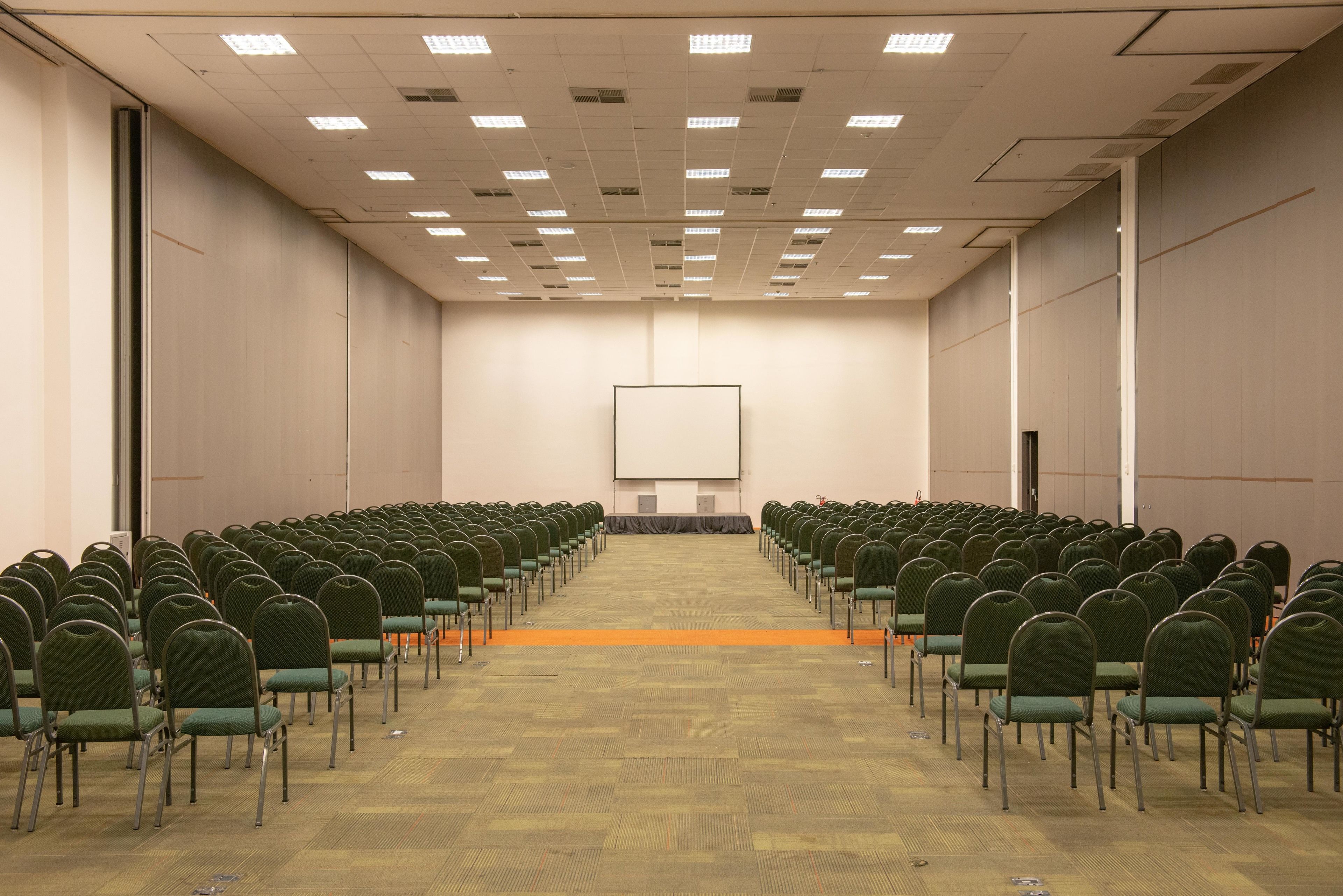 Une salle de conférence avec des rangées de chaises vertes, un sol recouvert de moquette et un grand écran blanc à l’avant.