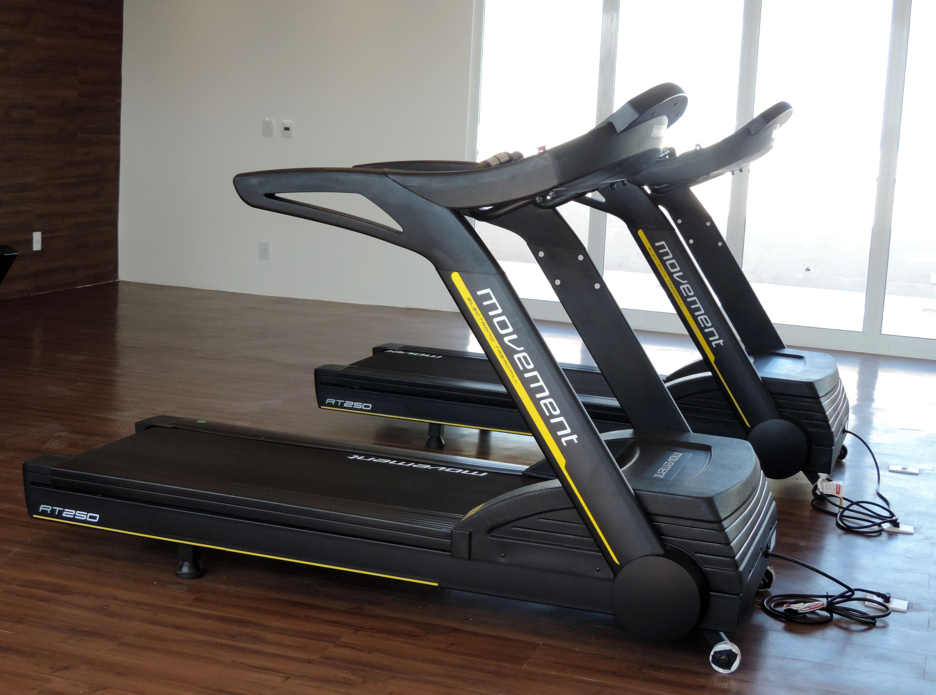 Two treadmill exercise machines with the brand name 'Movement' and model 'RT250' in a room with wooden flooring and a large window.
