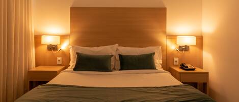 A hotel room with a large bed, two bedside lamps, a nightstand with a telephone, and a wooden headboard.