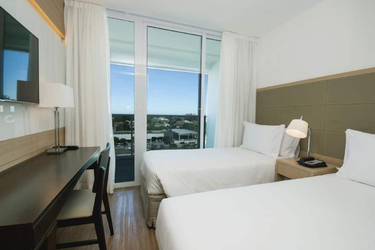A hotel room with two beds, a desk, a chair, a TV, and a view of the city.