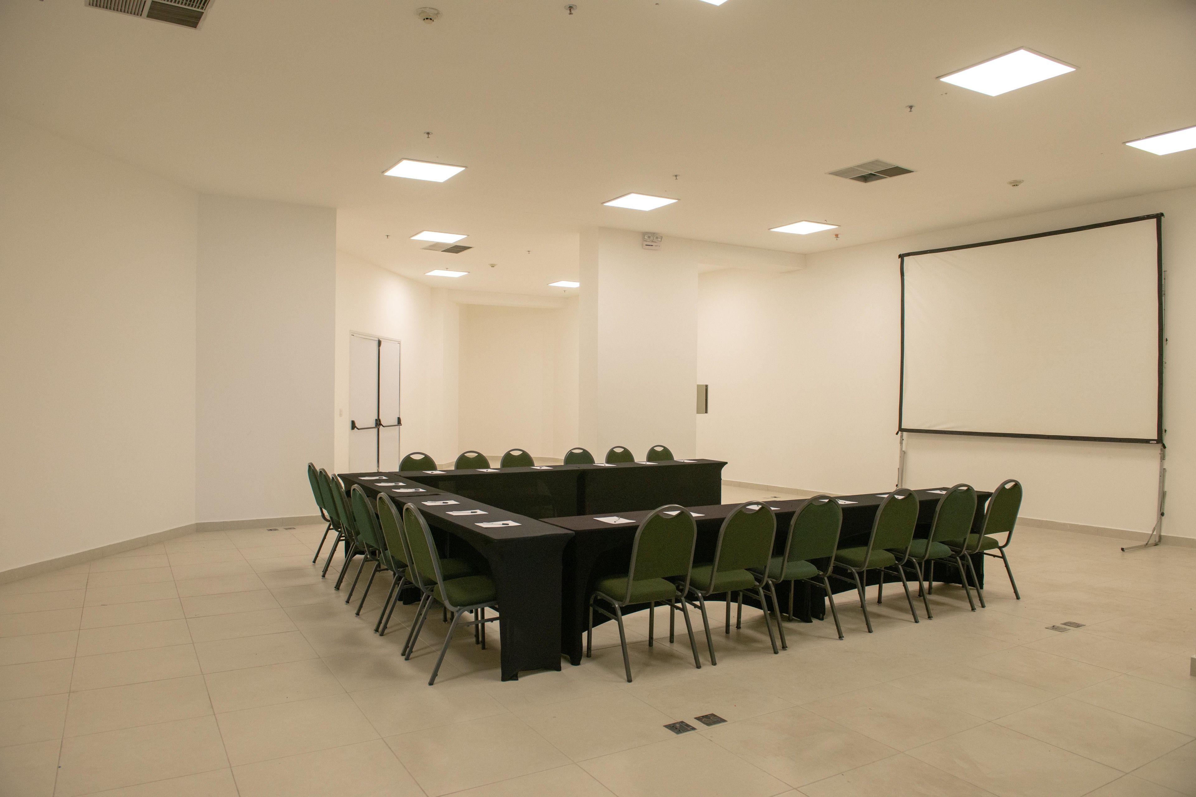 Un salón de conferencias con una larga mesa, sillas verdes, una pantalla de proyector y una pizarra.