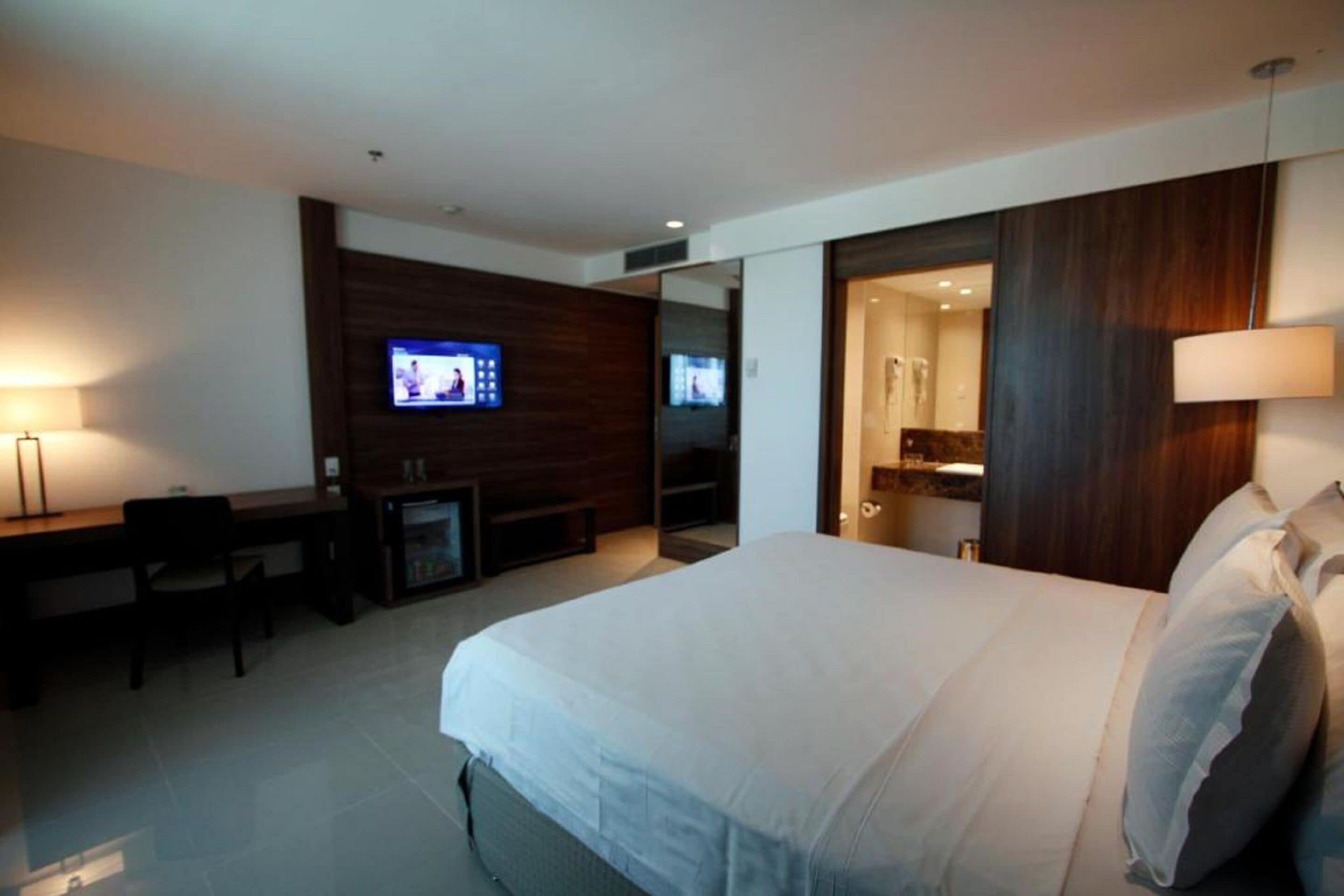 A modern hotel room with a large bed, a desk with a chair, a television, and a bathroom visible through an open door.