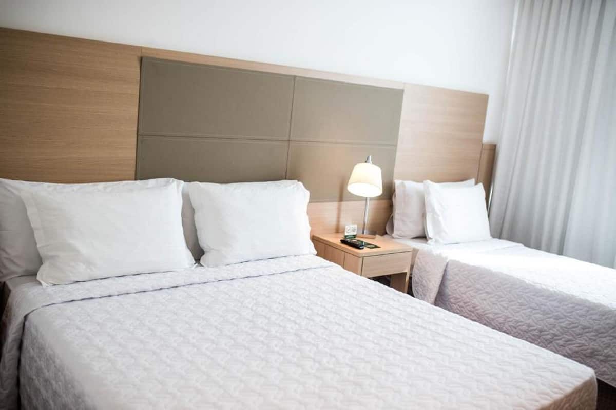 A hotel room with two beds, a wooden headboard, a nightstand with a lamp, and a window with curtains.