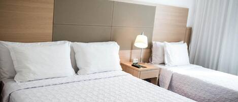 Habitación de hotel con dos camas, cabecero de madera, mesita de noche con lámpara y ventana con cortinas.