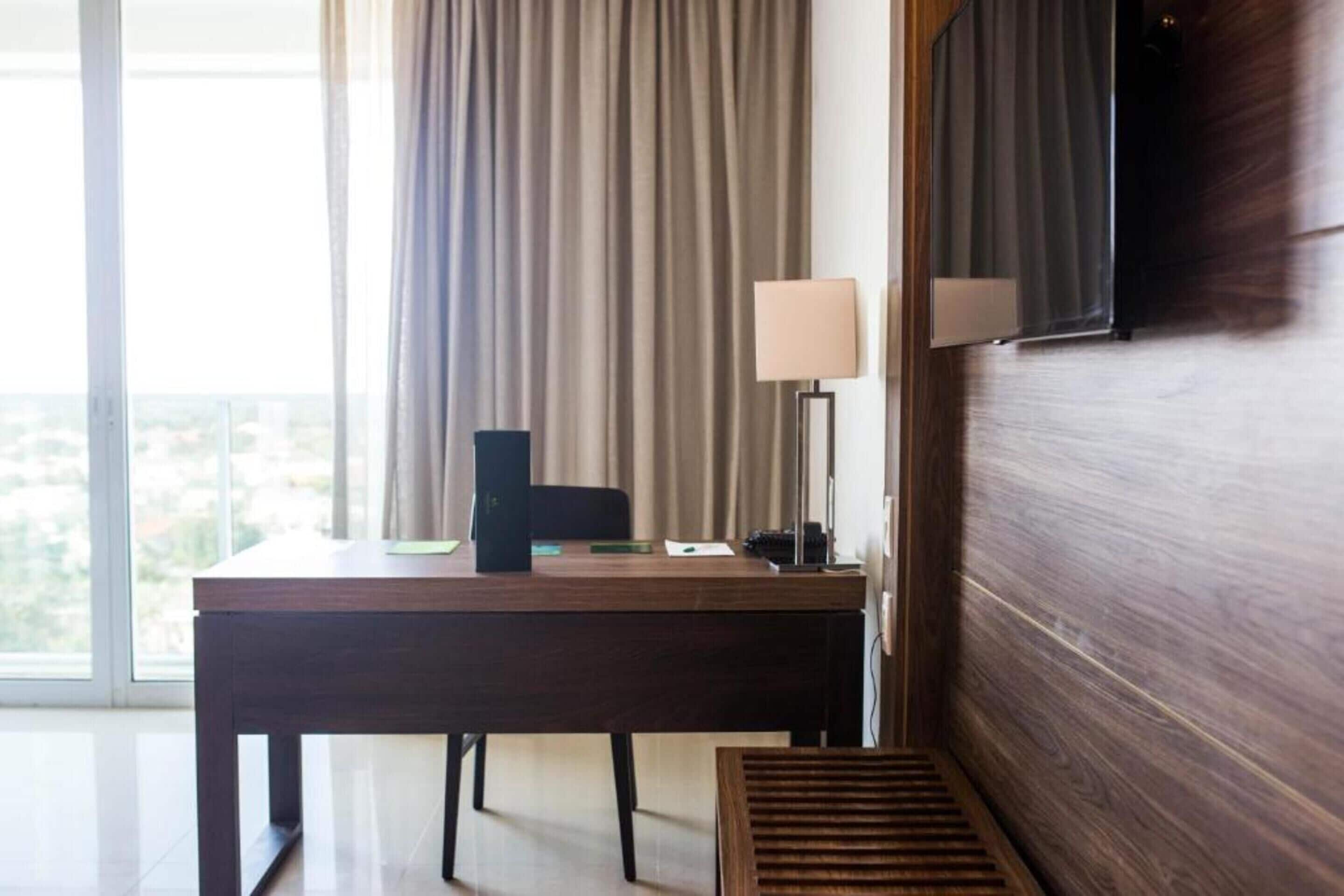 A hotel room with a wooden desk, a chair, a TV mounted on the wall, and a window with curtains.