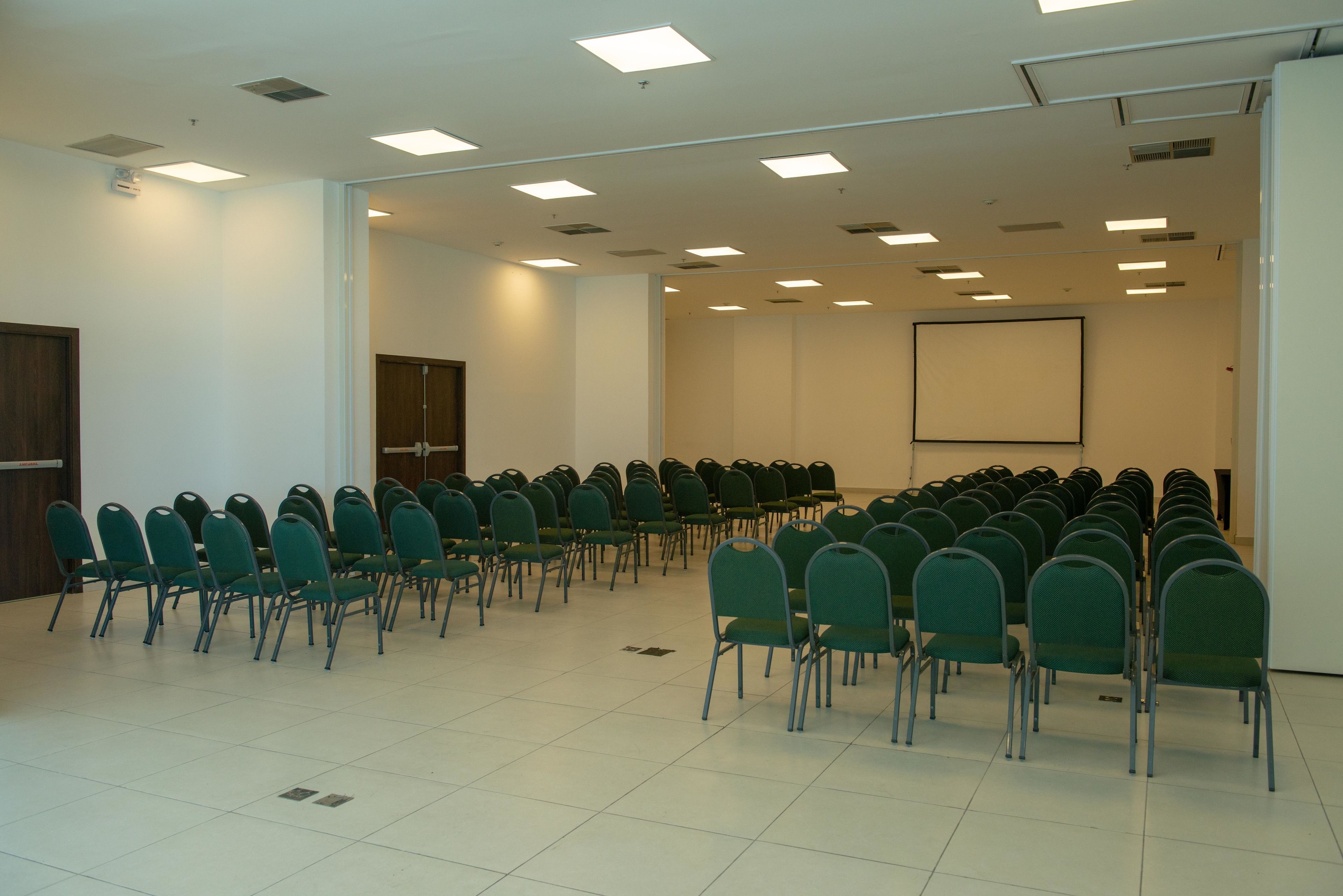 Un salón de conferencias con filas de sillas verdes, una pantalla de proyector y una pizarra.