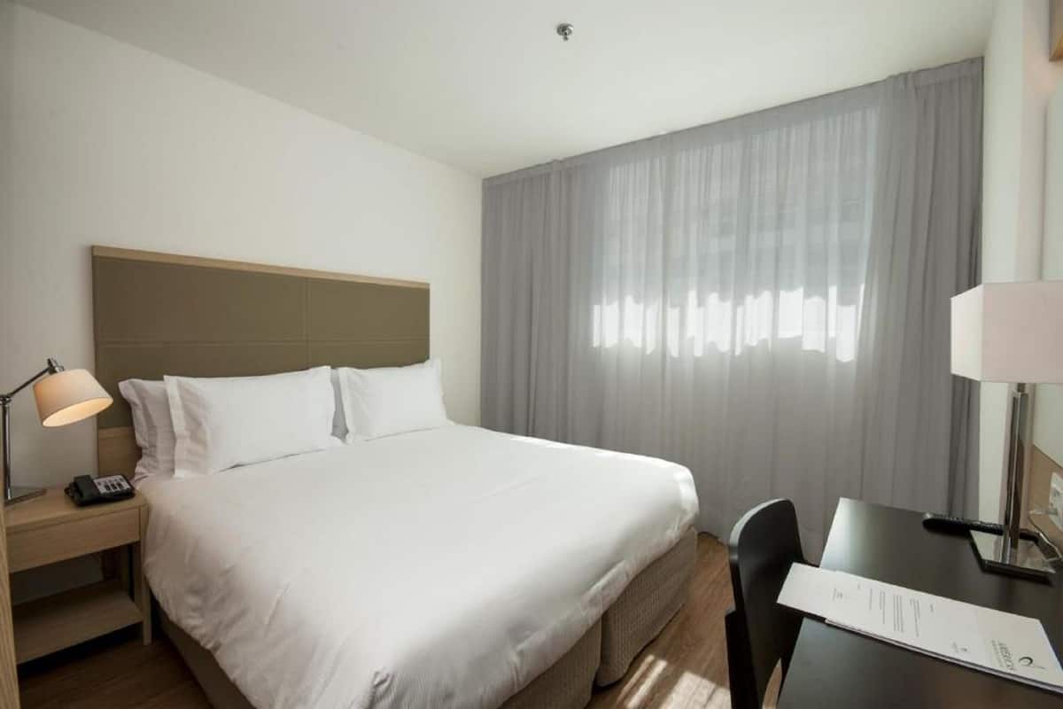 A hotel room with a bed, bedside table, lamp, and a desk with a chair.