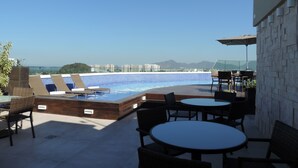 Uma área de piscina no terraço com espreguiçadeiras e mesas, vista para a paisagem urbana e céu claro.