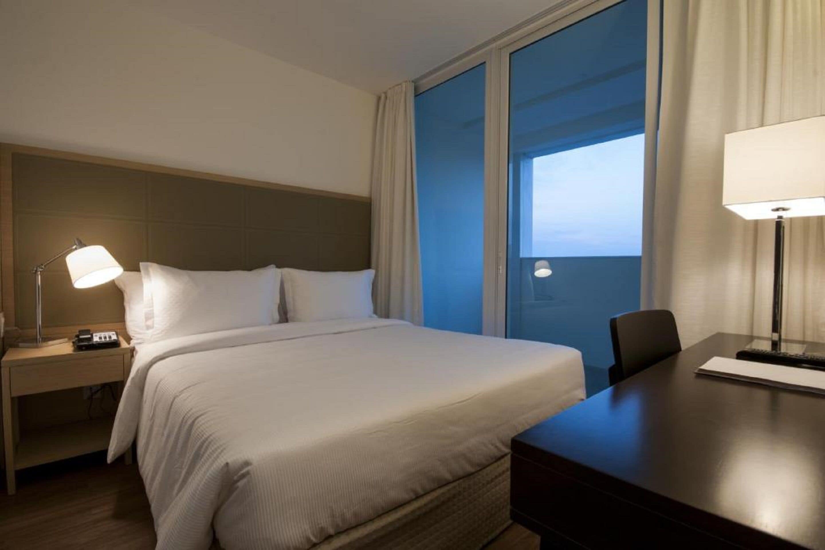 Una habitación de hotel con cama, mesitas de noche, un escritorio y un ventanal con cortinas.