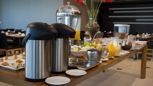 Uma mesa de buffet com diversos alimentos e bebidas, incluindo uma cafeteira, uma cesta de frutas e um vaso com flores.