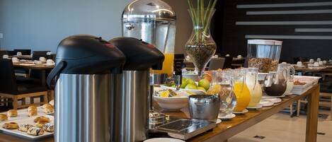 A buffet setup with various food items and beverages, including a coffee maker, a fruit bowl, and a vase with flowers.