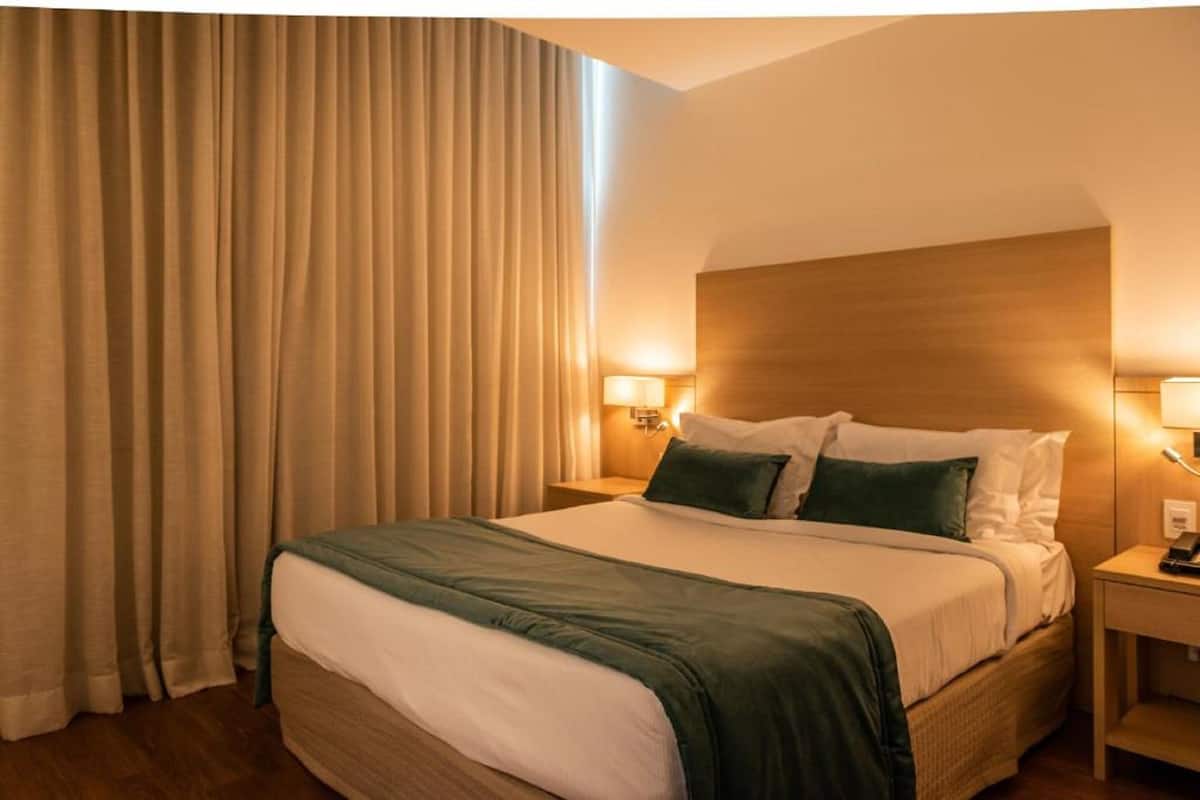 A hotel room with a large bed, two bedside lamps, a wooden headboard, and a curtain on the window.