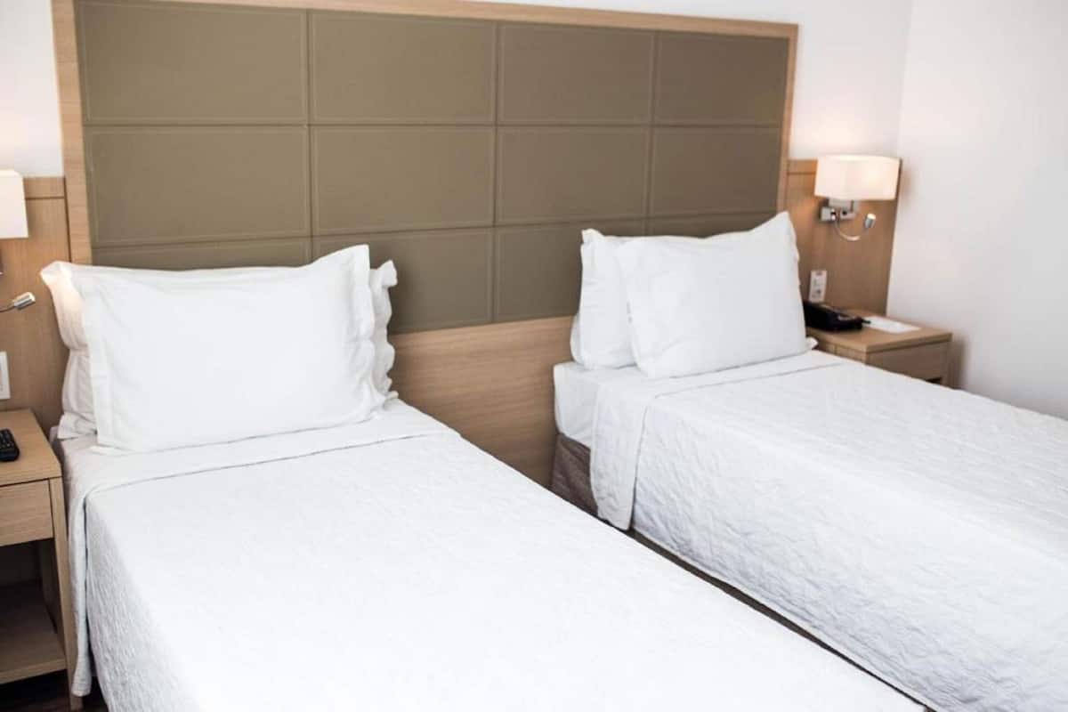 A hotel room with two single beds, a wooden headboard, bedside lamps, a nightstand, and a small drawer.
