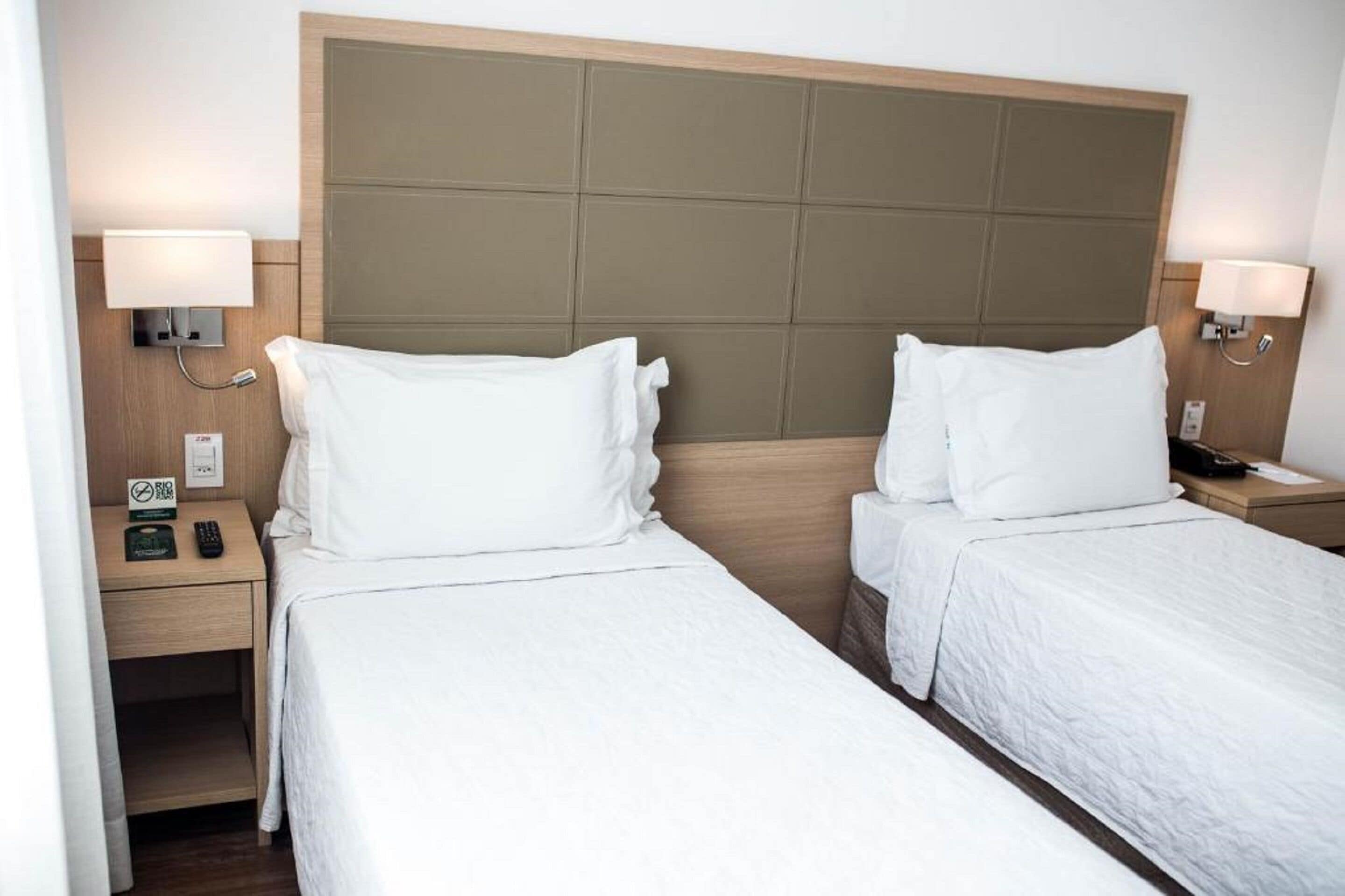 A hotel room with two single beds, a wooden headboard, bedside lamps, a nightstand with a phone and a clock, and a wall-mounted light switch.