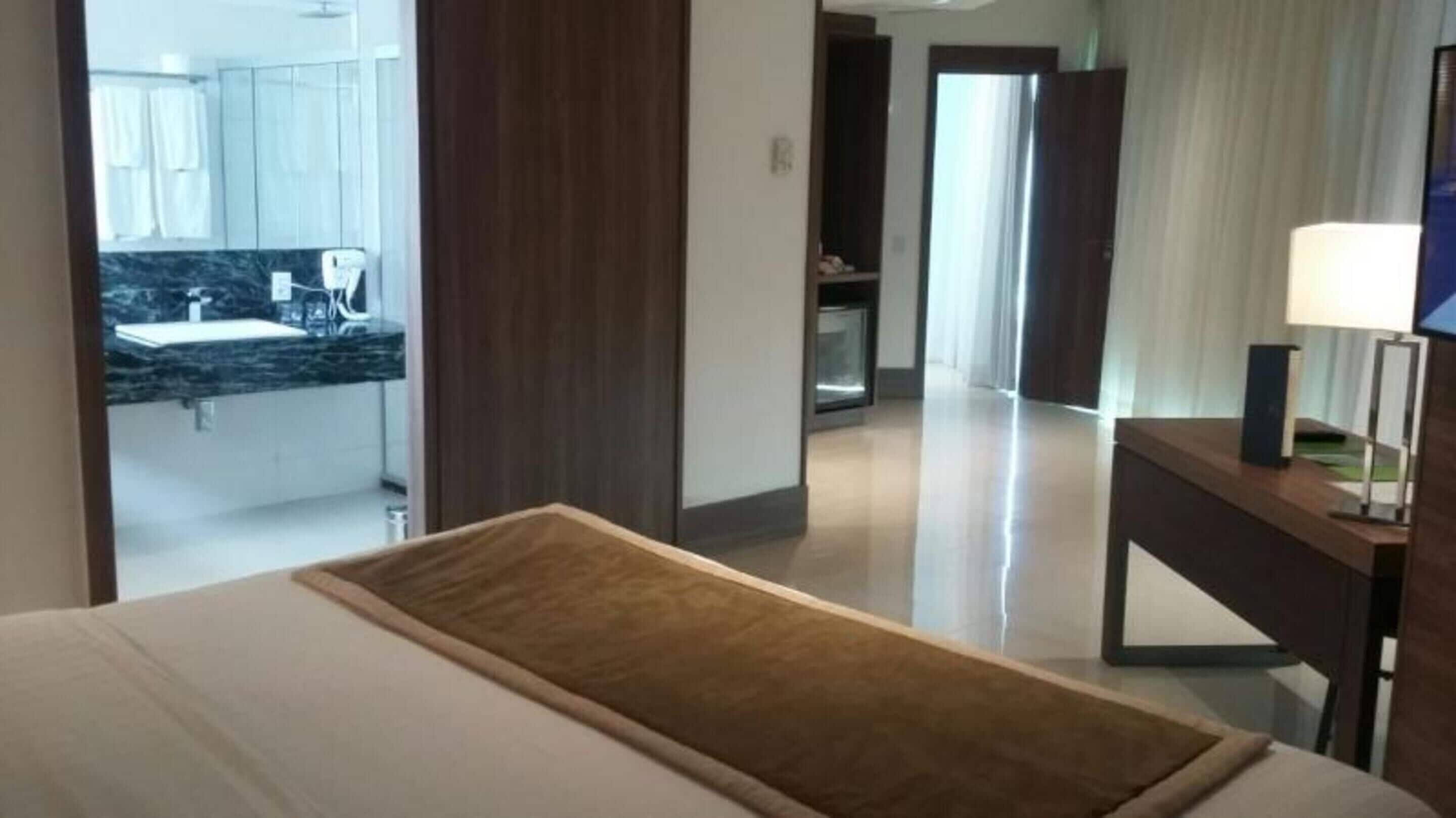 Habitación de hotel con cama, mesita de noche de madera, escritorio con lámpara y vistas al baño y la cocina.