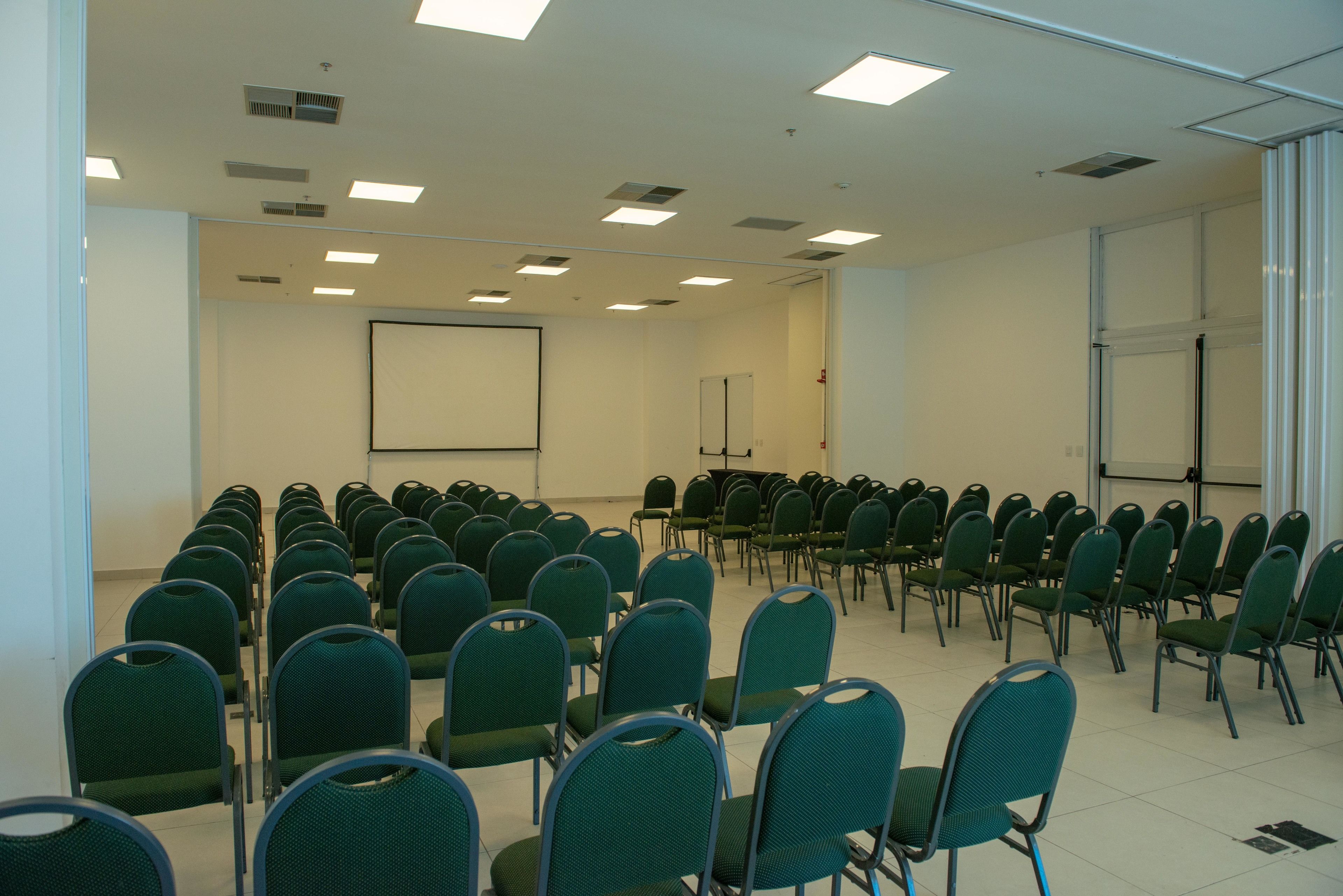 A conference room with rows of green chairs, a whiteboard, and a projector screen.