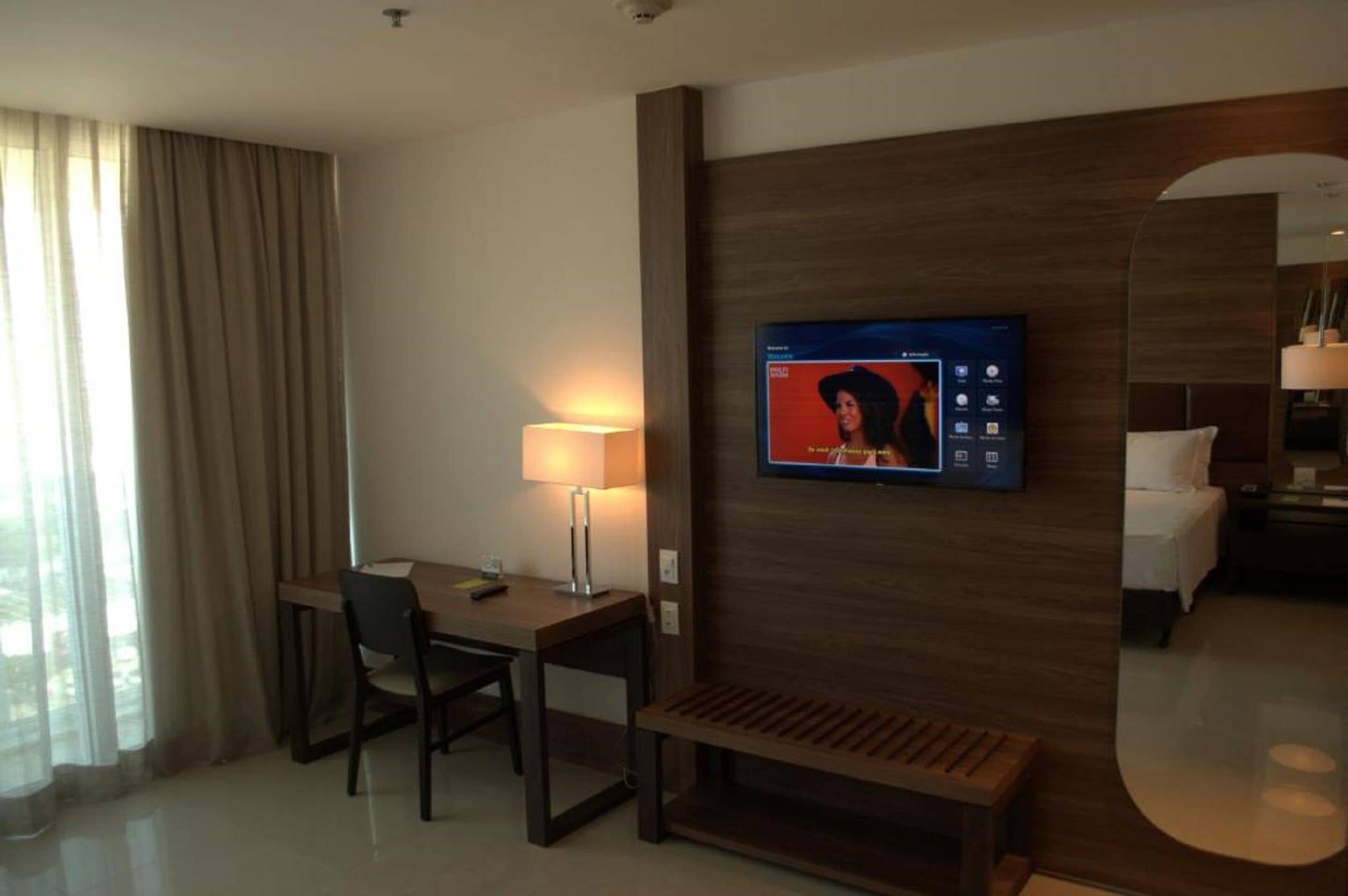 A hotel room with a wooden desk, a chair, a TV mounted on the wall, a bed, and a bedside table.