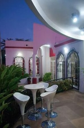 Un patio al aire libre con una mesa redonda y cuatro sillas blancas, rodeado de plantas en macetas y un edificio rosa con ventanas arqueadas.