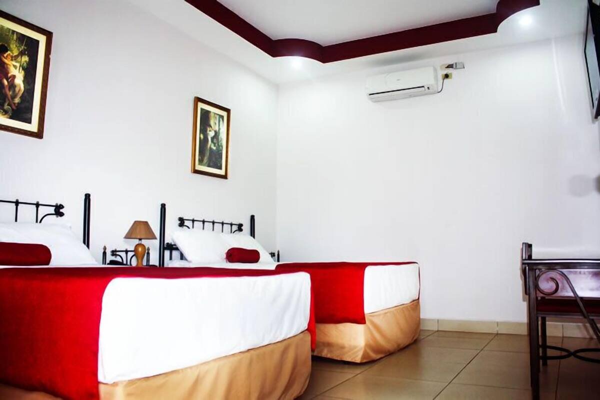 A hotel room with two beds, each with red and white bedding, a small table with a lamp, and a TV on the wall.