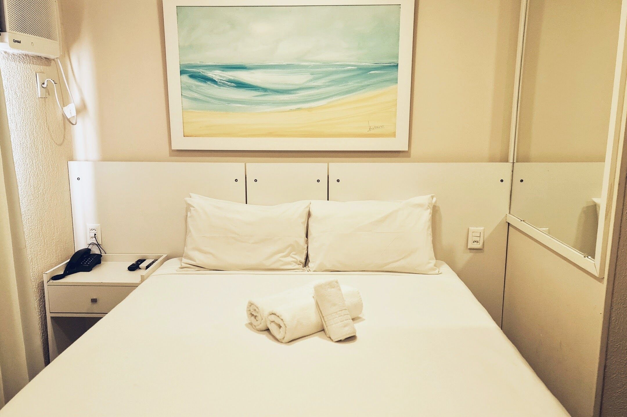 A hotel room with a bed, a painting of a beach, a bedside table with a phone, and a wall-mounted mirror.