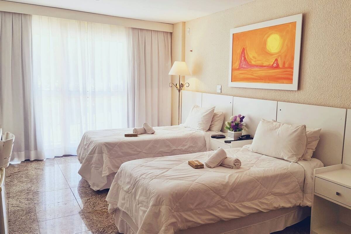 Habitación de hotel con dos camas, una mesita de noche con lámpara, un cuadro en la pared y un ventanal con cortinas.