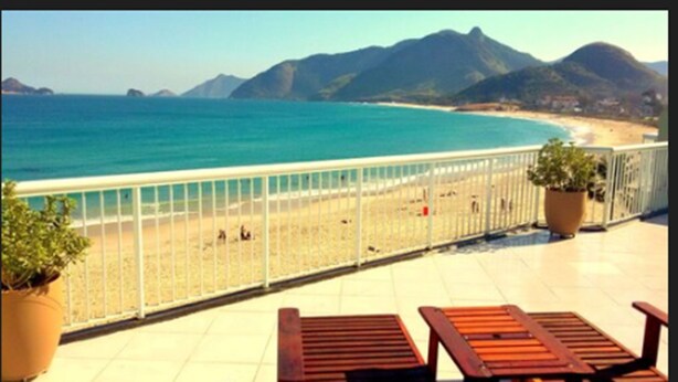 Een balkon met uitzicht op strand en bergen. Twee potplanten en twee houten stoelen.