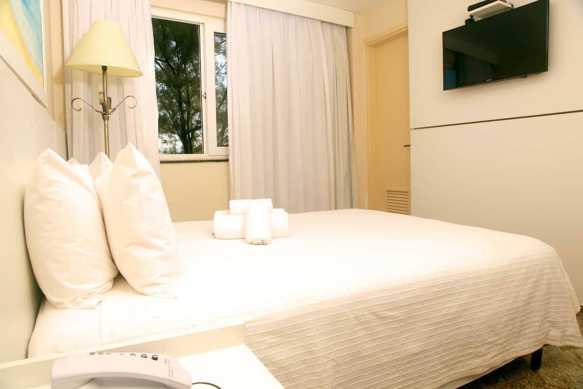 Una cama bien hecha con almohadas y toallas enrolladas, una lámpara de noche y un televisor de pantalla plana montado en la pared.