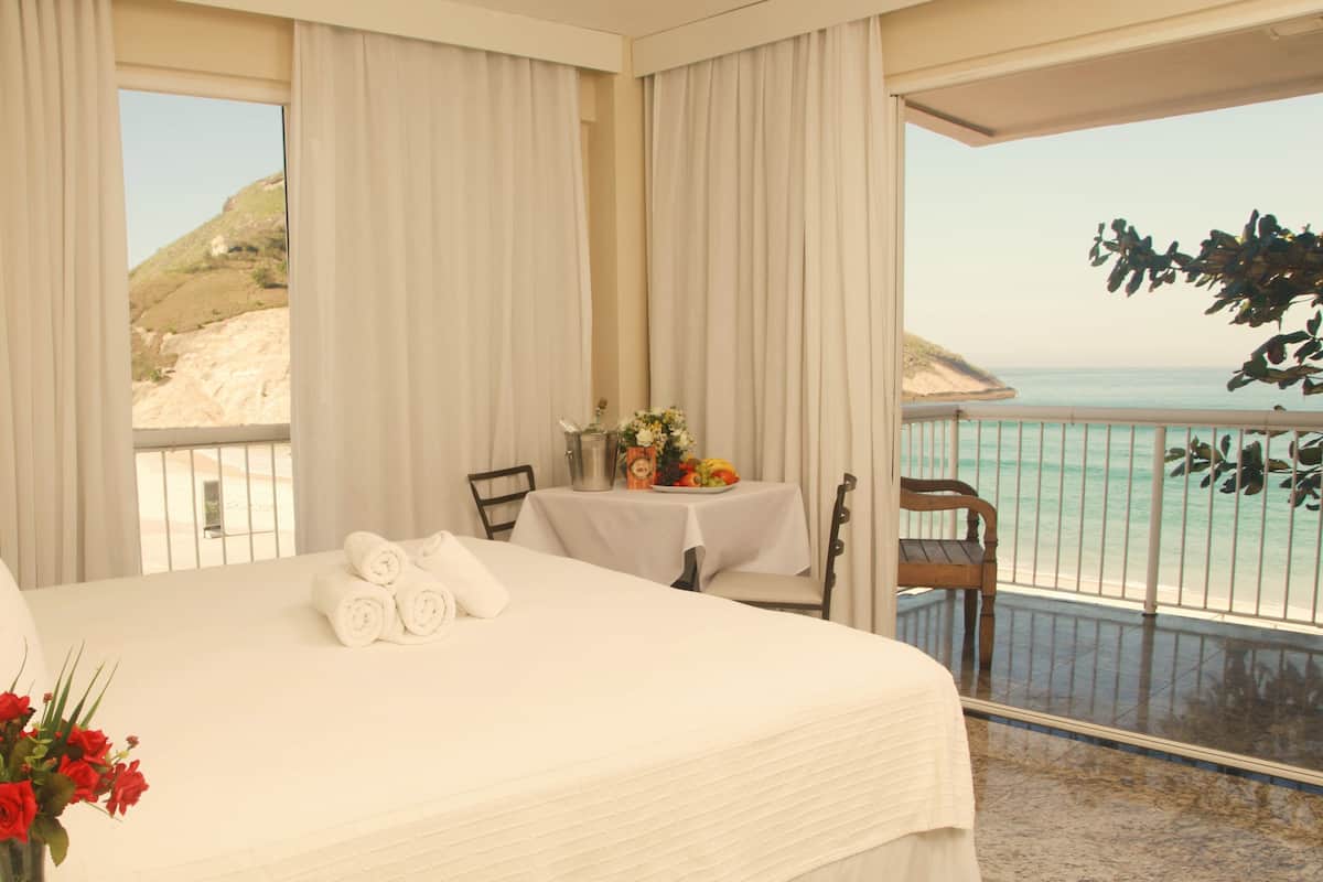 Un cuarto de hotel con cama, una mesa con un jarrón de flores y vista al mar.
