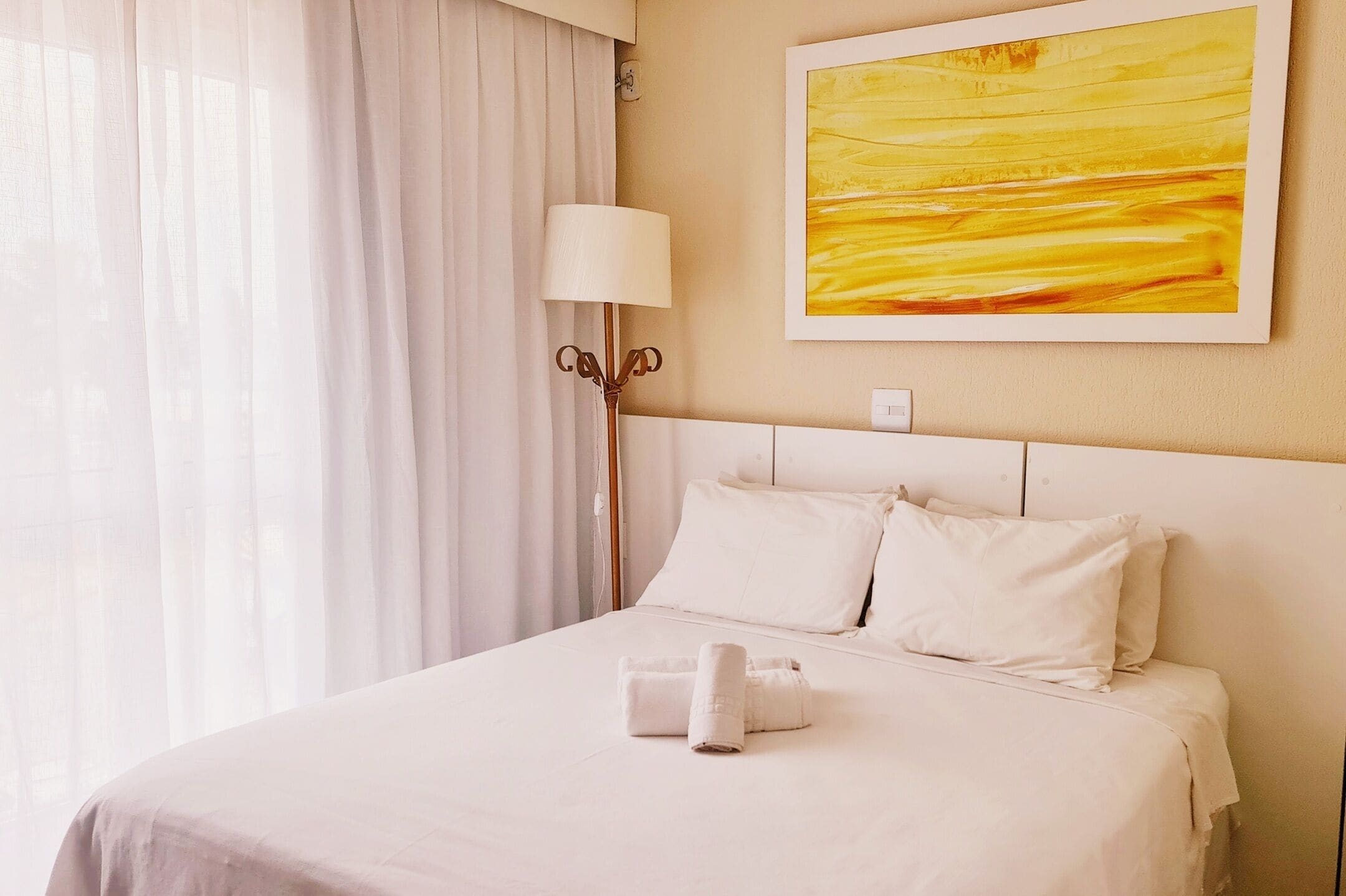 Une chambre d’hôtel avec un lit, des rideaux blancs, une lampe et un tableau au mur.
