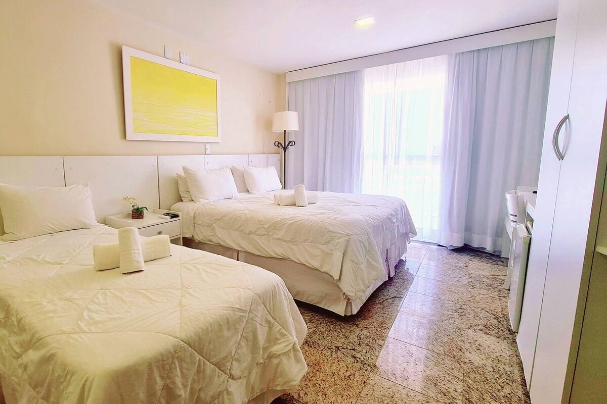 Habitación de hotel con dos camas, un ventanal con cortinas, una lámpara de noche y un cuadro en la pared.