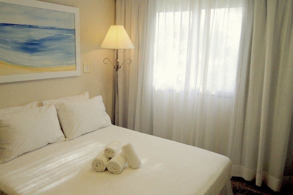 Uma cama bem arrumada com lençóis e toalhas brancas, uma luminária e um quadro na parede.