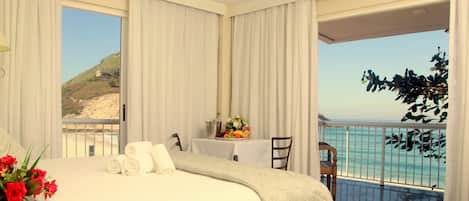 A hotel room with a large bed, a dining table with chairs, a balcony with a view of the sea, and a vase with flowers on the bed.