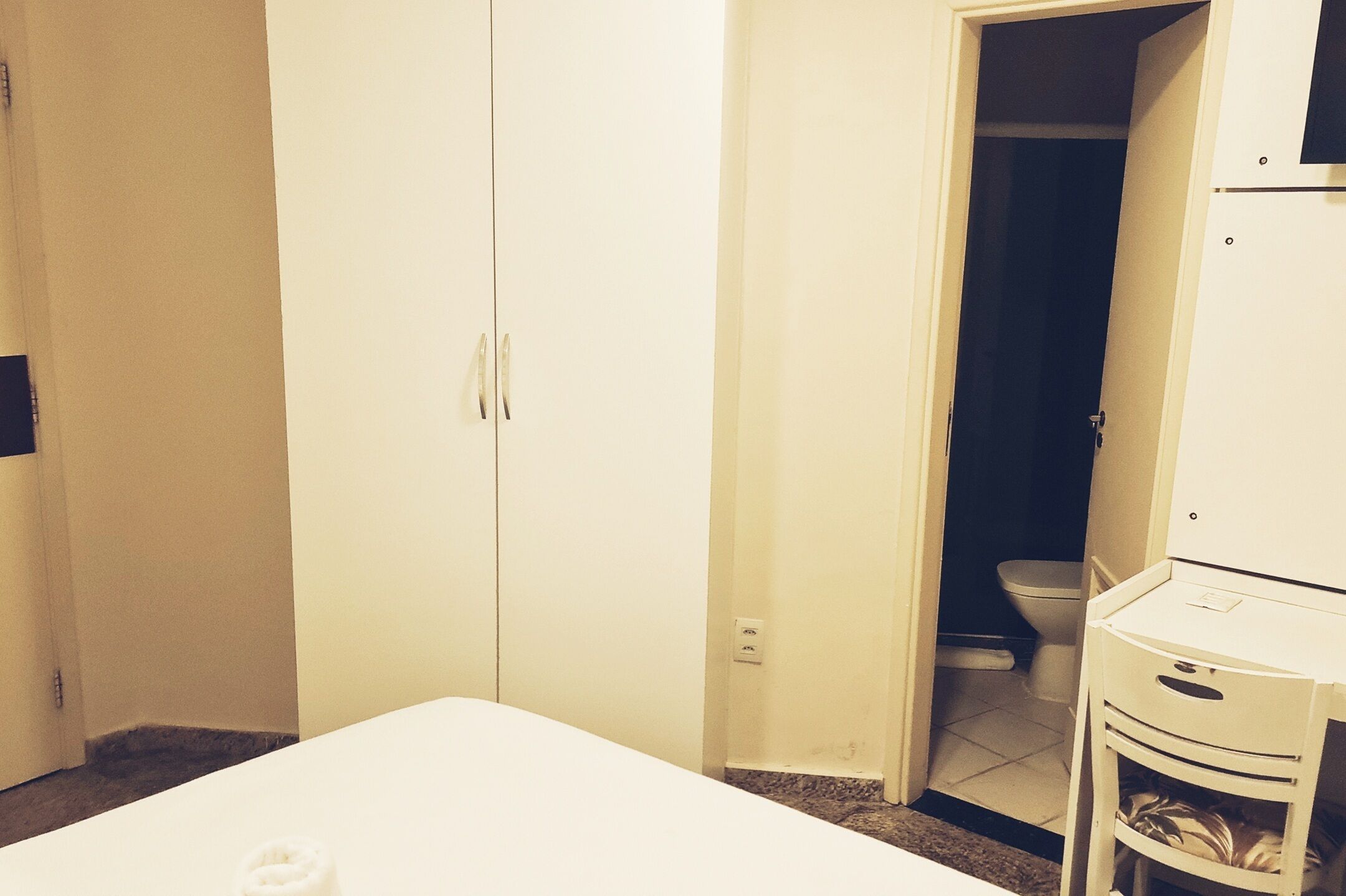 A room with a white bed, a white wardrobe, and a white cabinet. There is a door leading to a bathroom with a toilet.