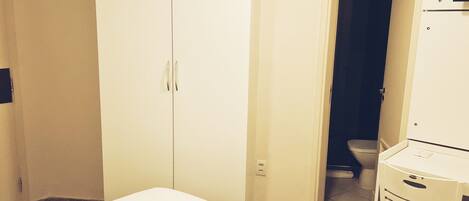 A room with a white bed, a white wardrobe, and a white cabinet. There is a door leading to a bathroom with a toilet.