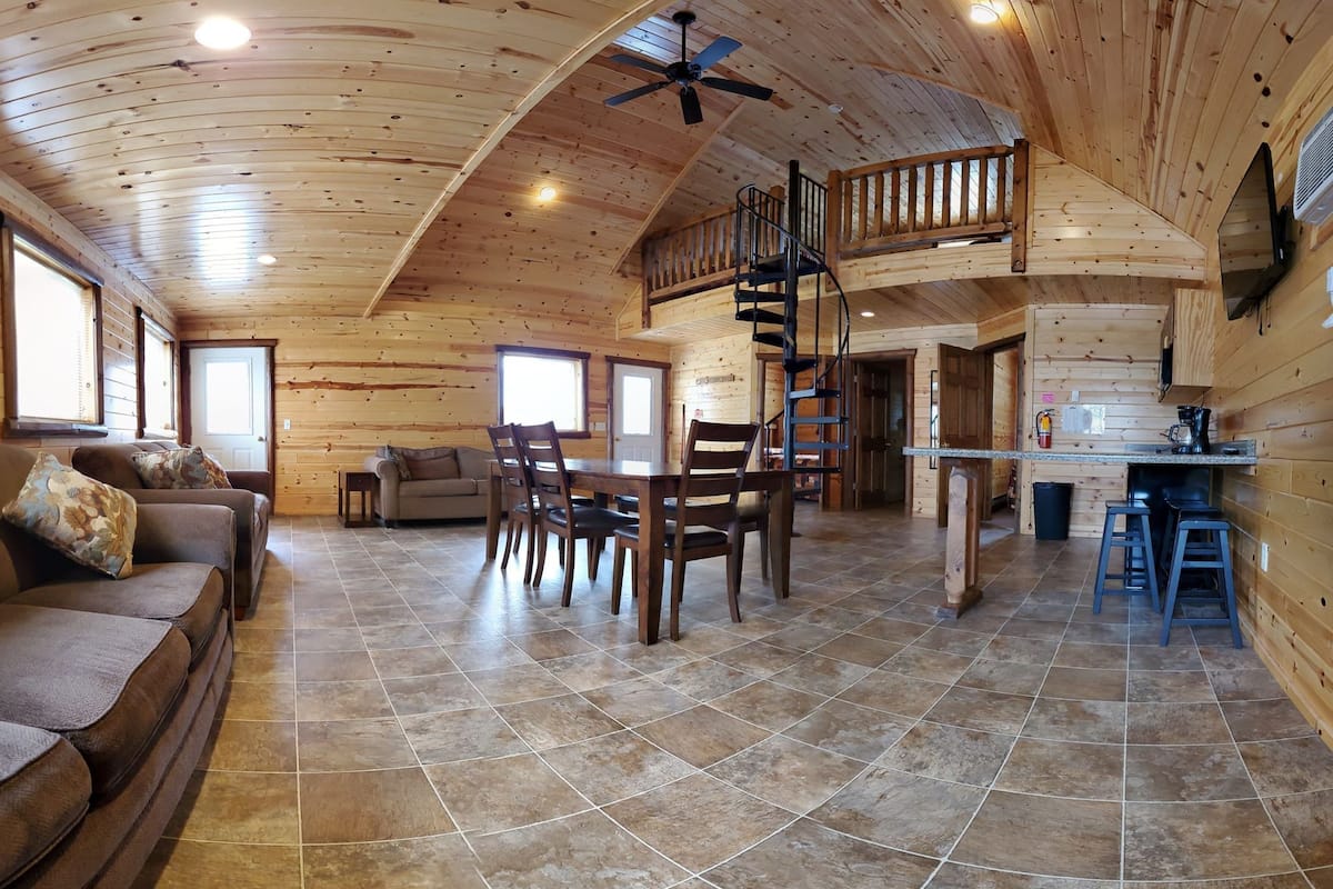 A spacious wooden interior with a dining area, a sofa, a staircase, and a kitchen area.