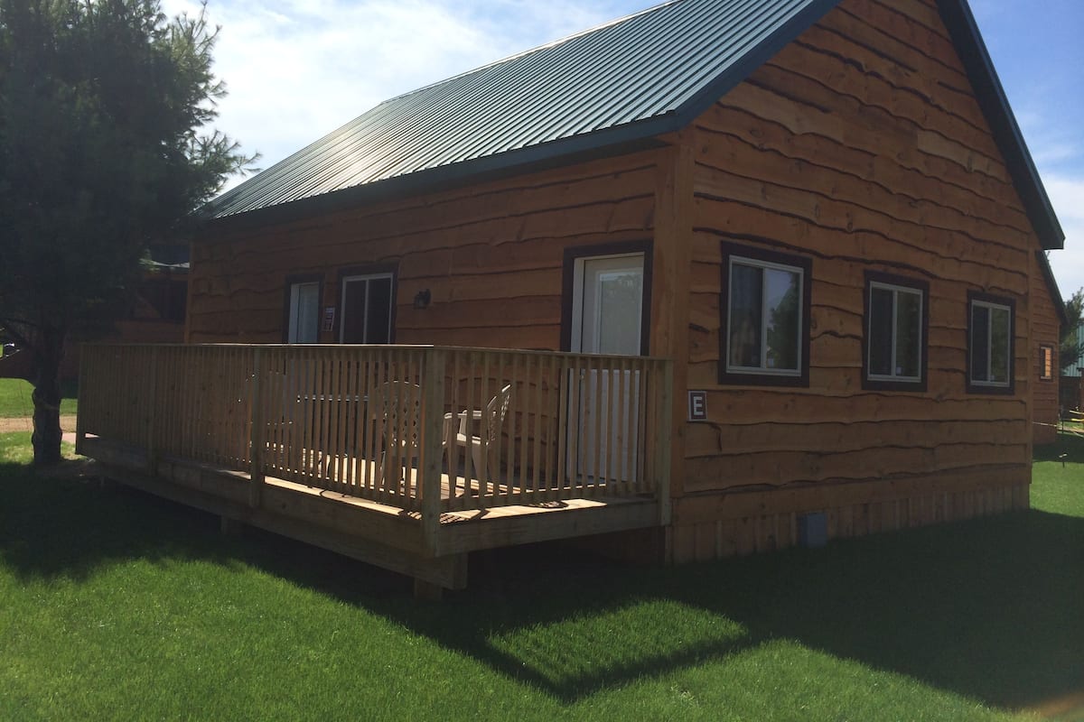 A wooden cabin with a metal roof, a deck with chairs, and a grassy yard.