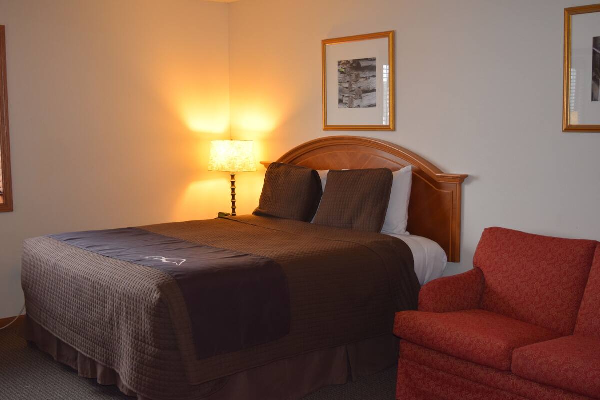 A hotel room with a bed, a nightstand with a lamp, a framed picture on the wall, and a red armchair.