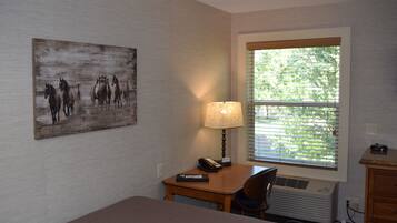 A hotel room with a bed, a desk with a lamp, a chair, a window with blinds, and a painting on the wall.