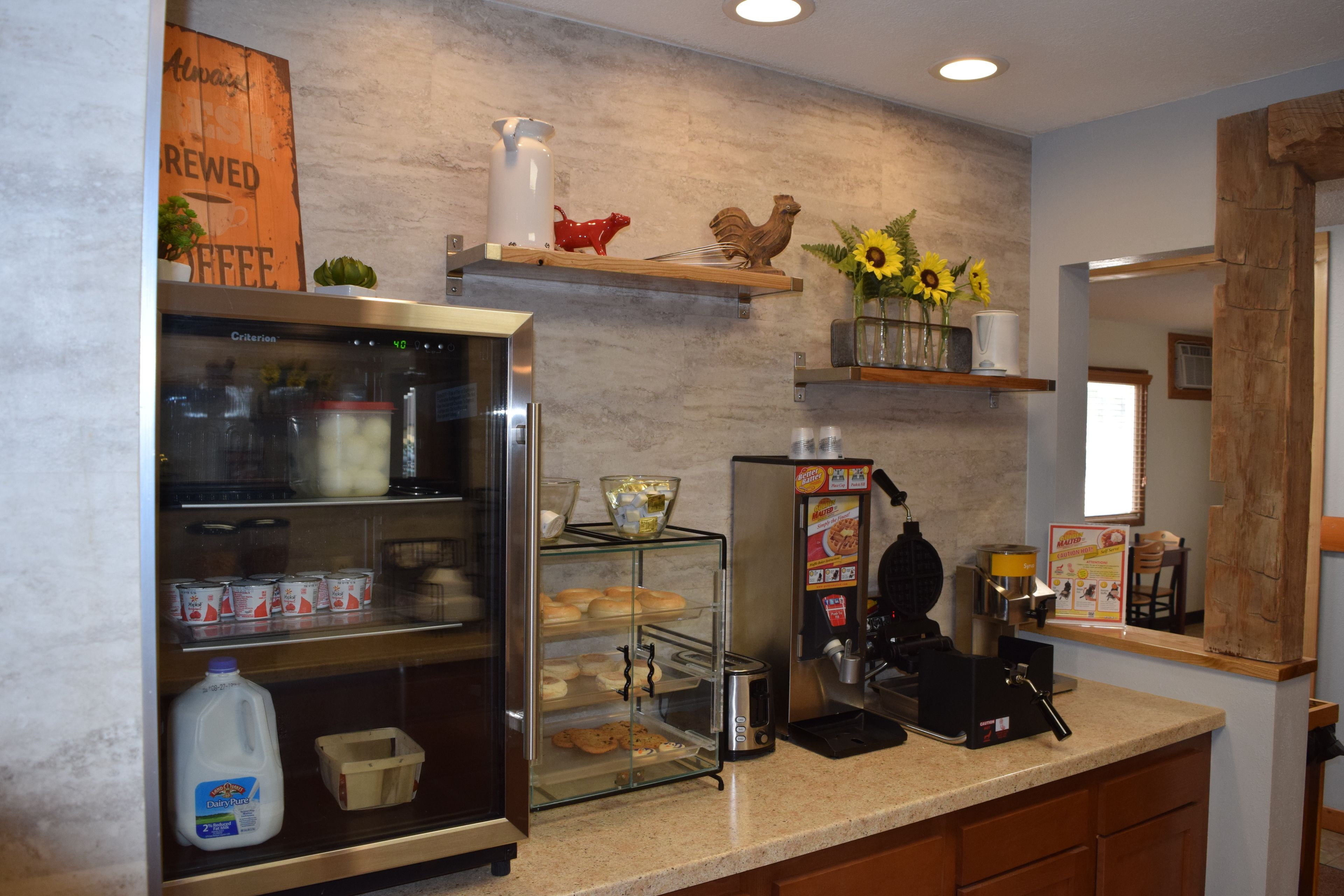 Un área de cocina con un refrigerador, una jarra de leche, una máquina de café y una vitrina con productos de repostería.