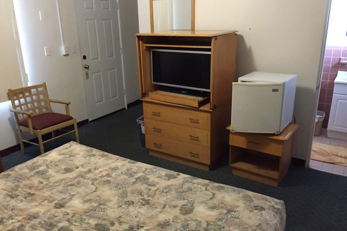 Une chambre d’hôtel comprenant un lit, un meuble en bois servant de support pour la télévision, un petit réfrigérateur, une chaise avec un coussin et une porte.
