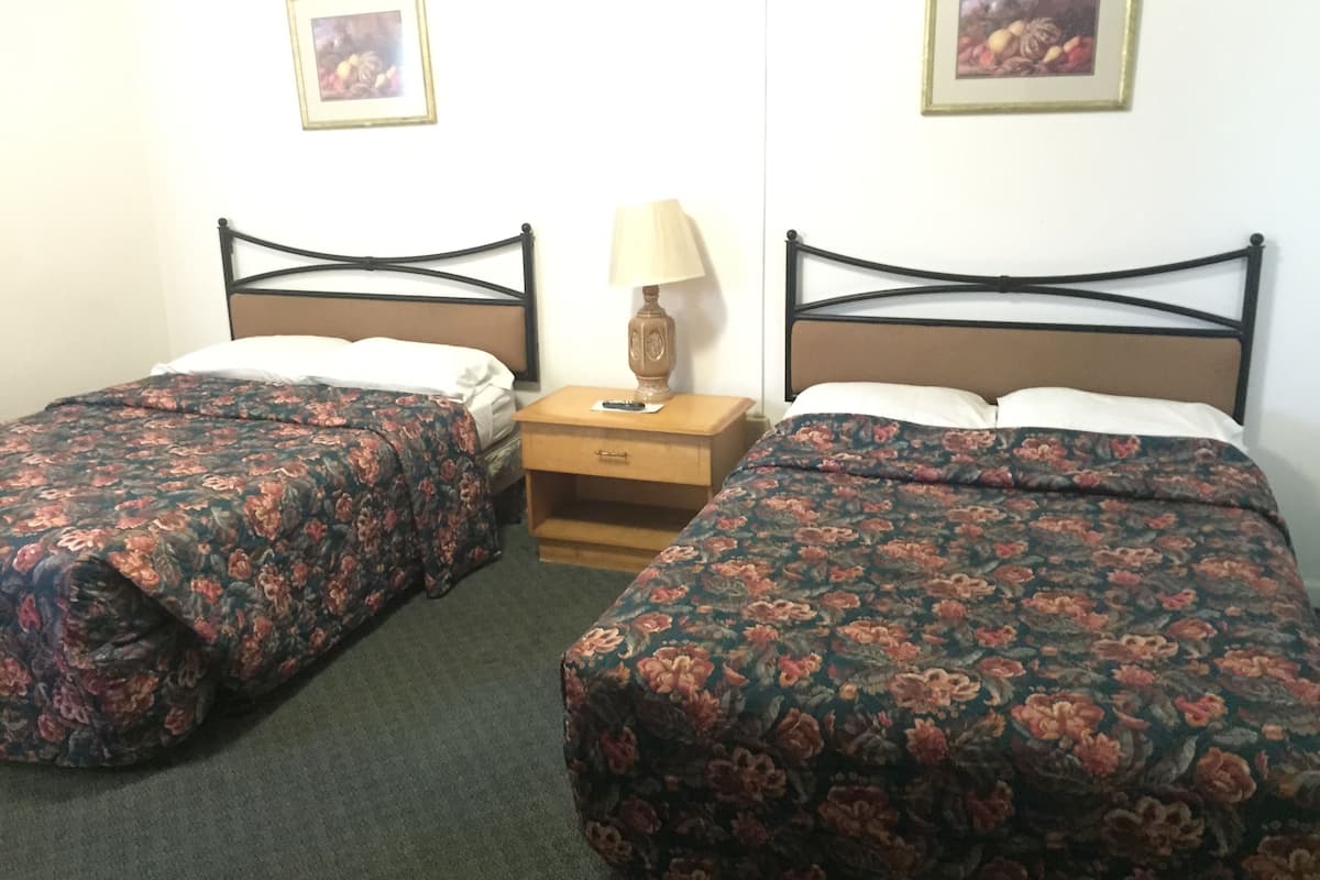 Une chambre avec deux lits simples, chacun recouvert d’une couvre-lit à motifs floraux, une table de chevet en bois avec une lampe, et des tableaux encadrés accrochés au mur.