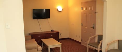 A hotel room with a flat-screen TV mounted on the wall, a wooden desk, a chair, a sofa, and a door.