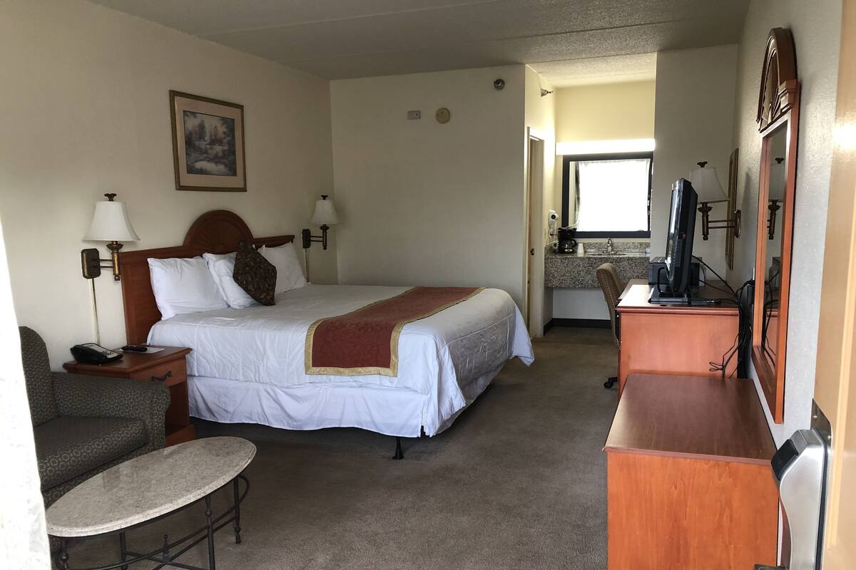 Quarto de hotel com cama, mesas de cabeceira, escrivaninha, cadeira, televisão e banheiro visível através de uma porta aberta.