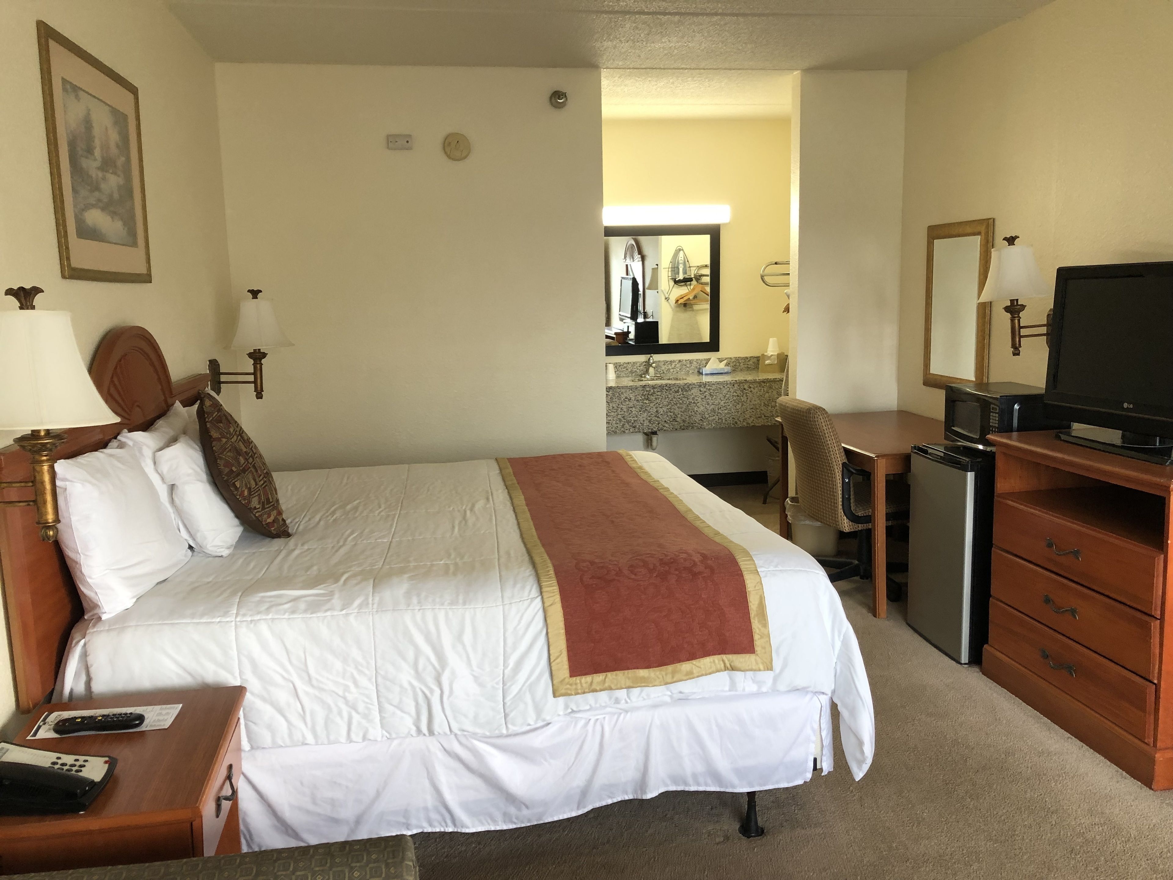 A hotel room with a bed, bedside tables, a desk with a chair, a television, a microwave, and a refrigerator.