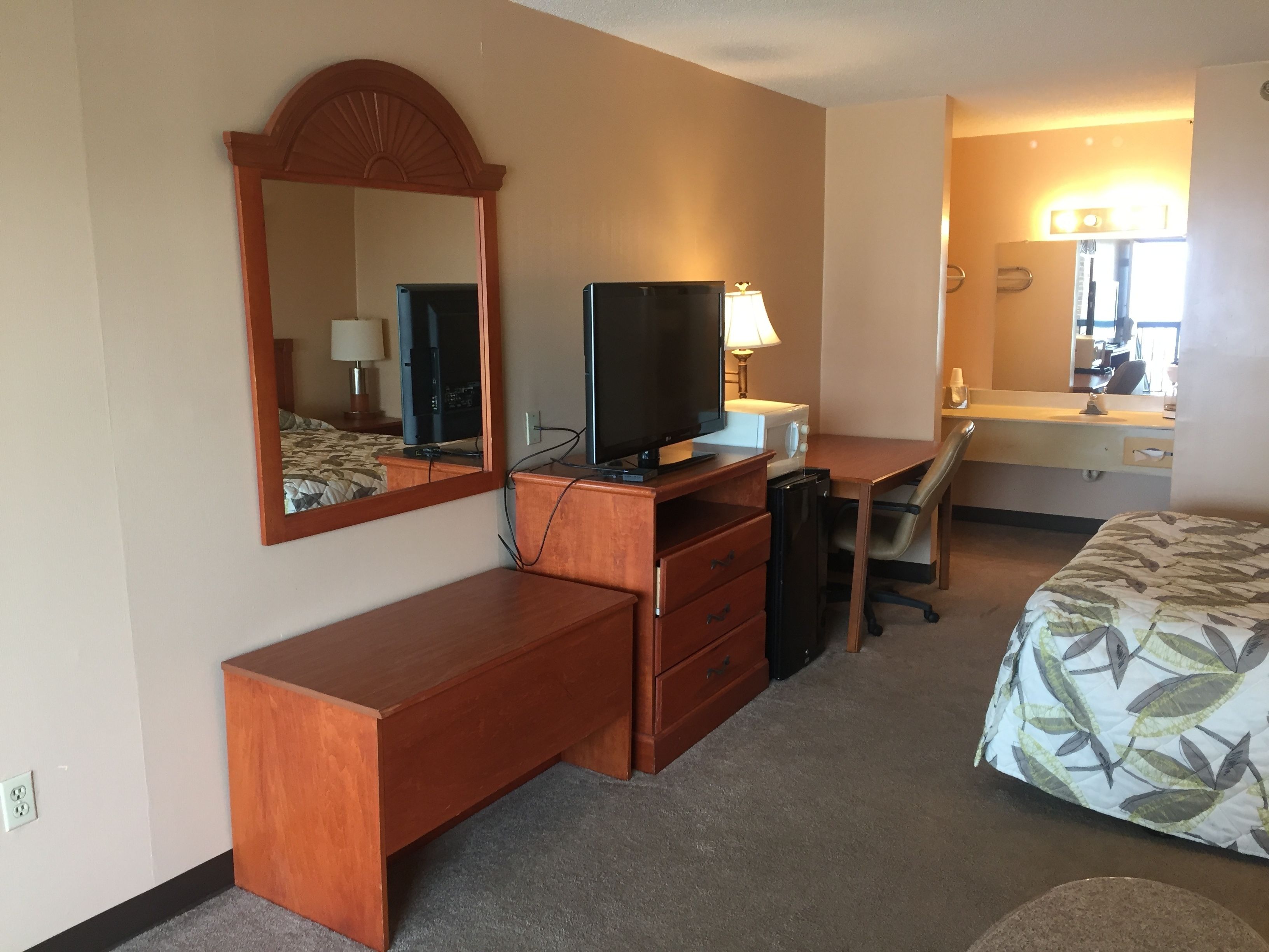 Une chambre d’hôtel avec un bureau en bois, une télévision, un miroir, un lit recouvert d’une couvre-lit à motifs de feuilles, et une salle de bain avec un miroir et un lavabo.