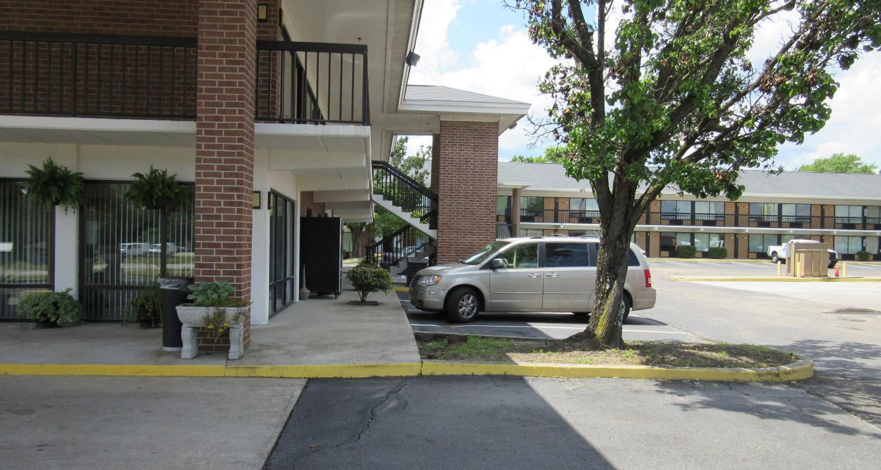 Un edificio con fachada de ladrillo, un balcón y un estacionamiento con un árbol y un coche aparcado.